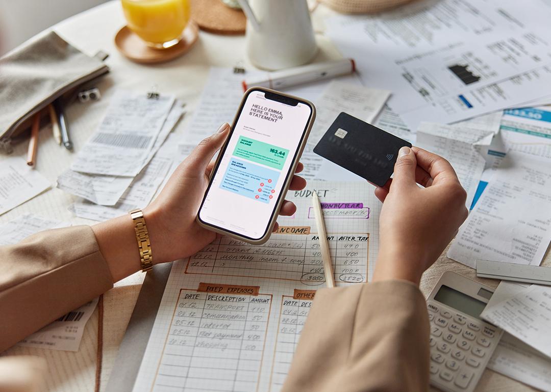 A woman organizes financial documents and is holding a credit card and a smartphone showing her statement.