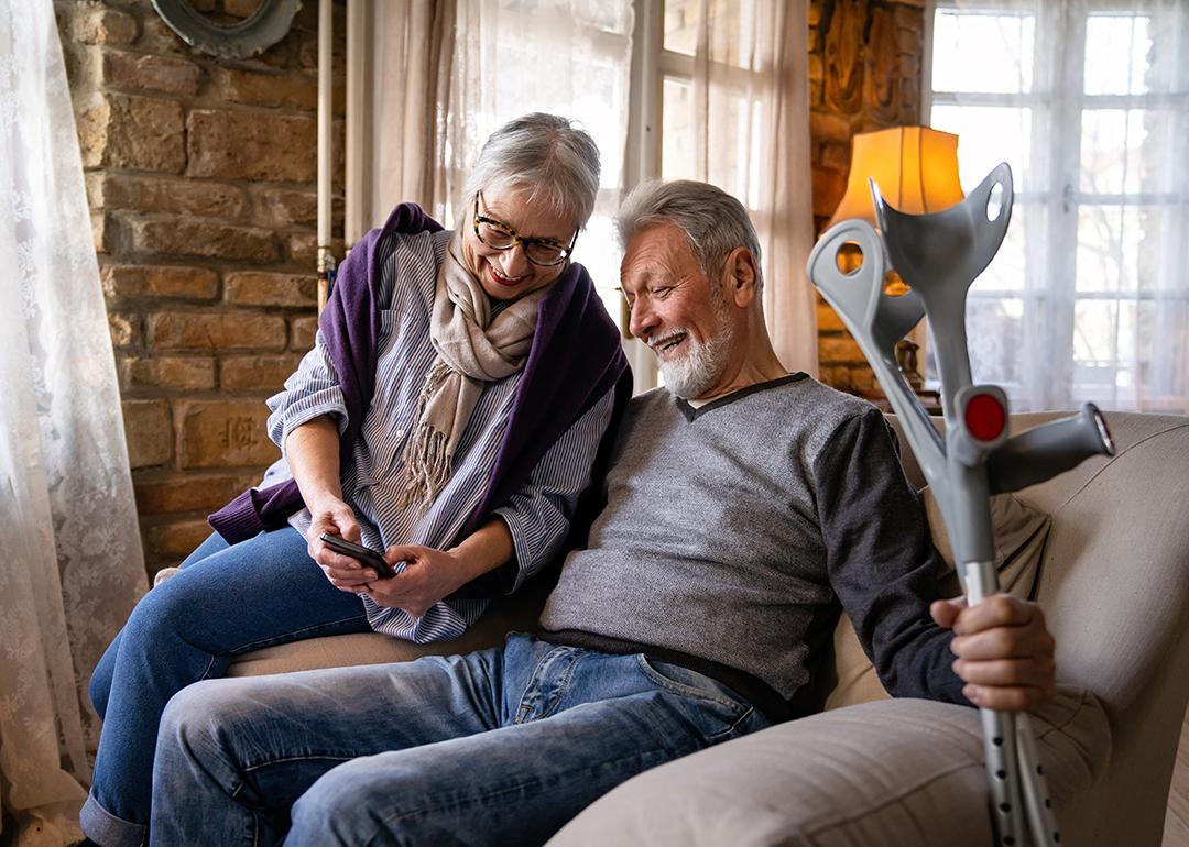 An elder couple where the wife shows her phone to her husband who is holding on to a pair of crutches while sitting on a sofa.