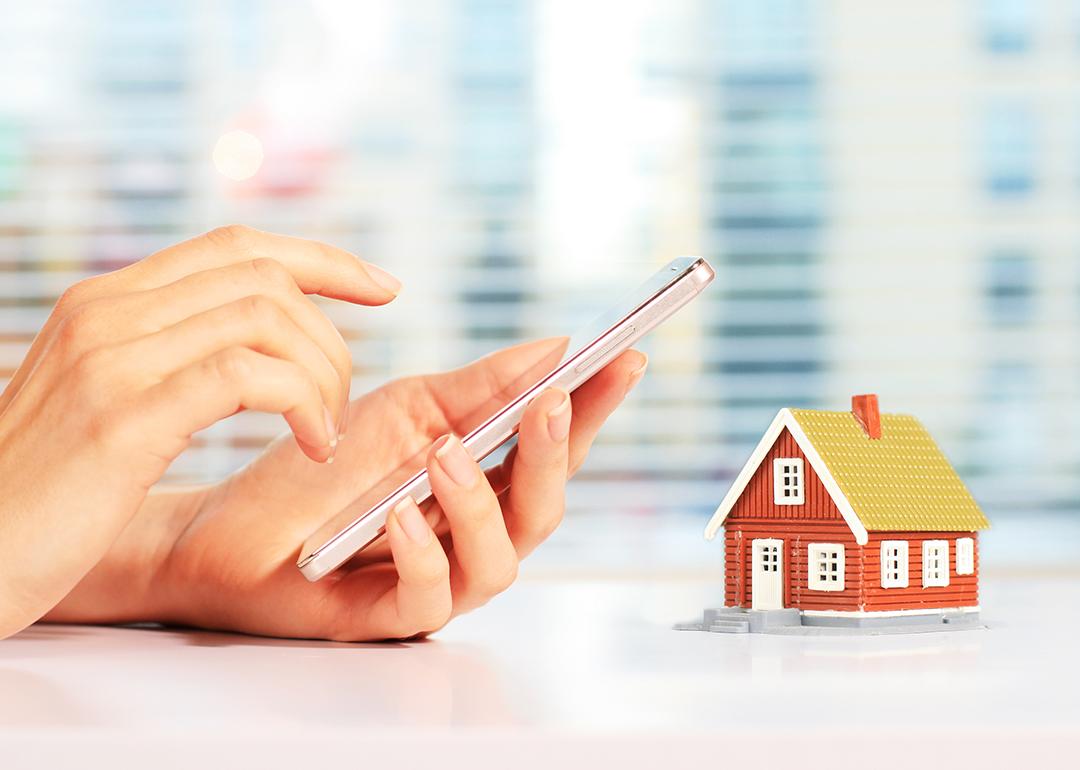 Person using smartphone and a figure of a house is on the same frame as a concept of paying rent digitally.