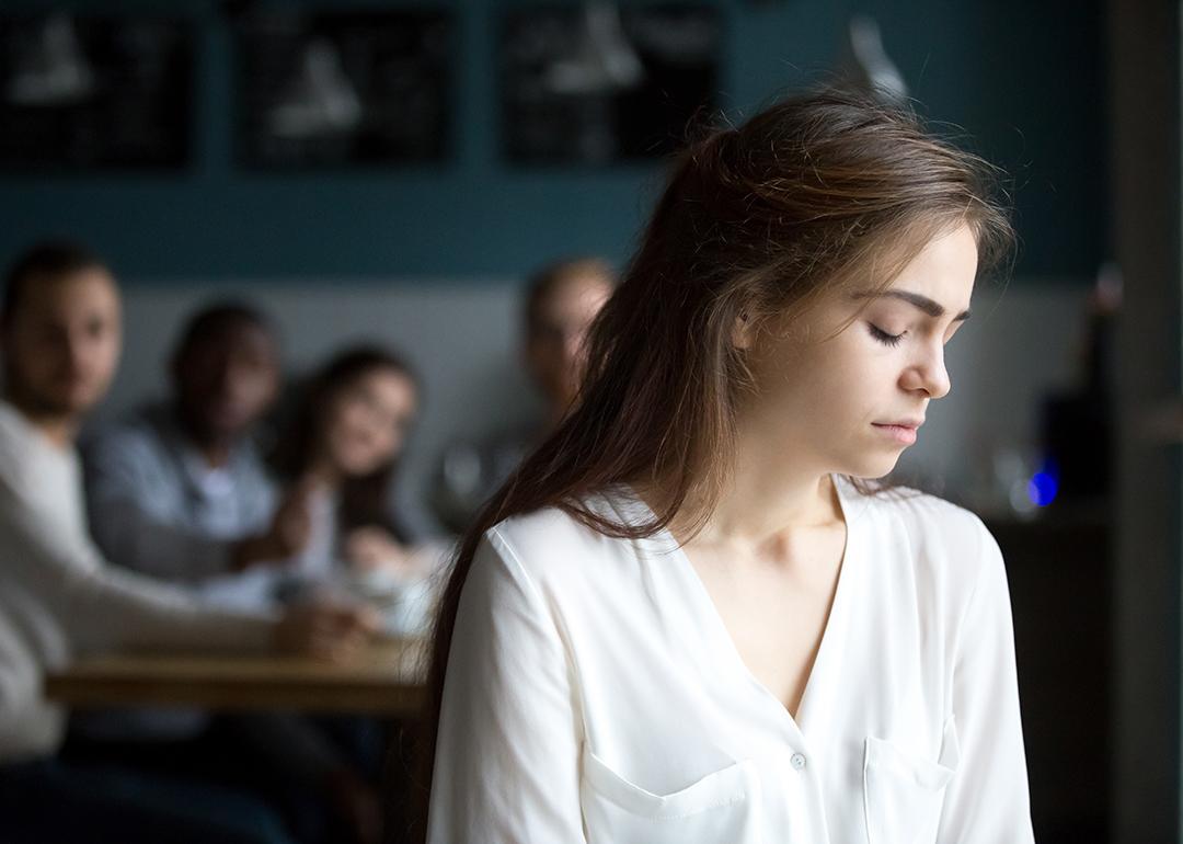 A sad young woman avoiding a group peers and sitting alone in a cafe.