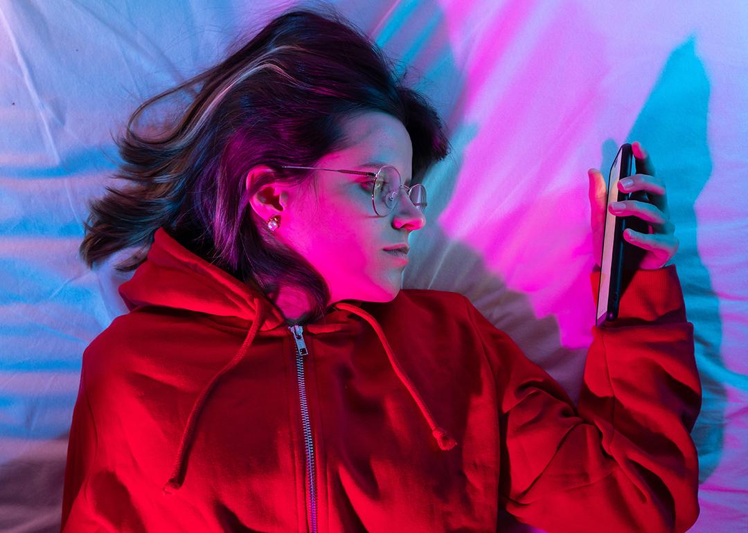 A young girl looking at her smartphone and doom-scrolling on the bed in her neon-lit room.