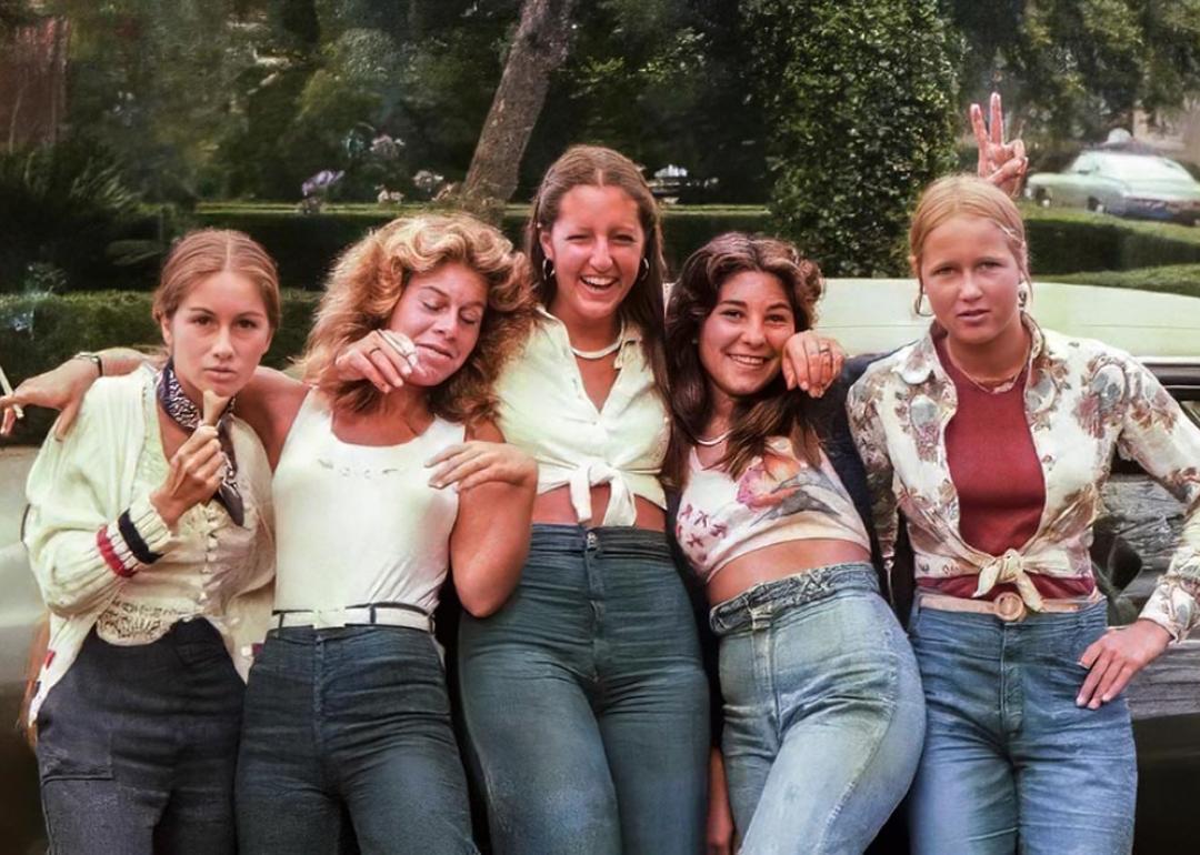 Color photo of five teenage girls leaning against a car on a tree-lined suburban street in San Diego in 1977. They wear high-waisted jeans and various casual tops including a tied blouse, a floral shirt, and a cardigan. One makes a peace sign and another rests her head on a friend's shoulder. Green hedges and parked cars are visible behind them.
