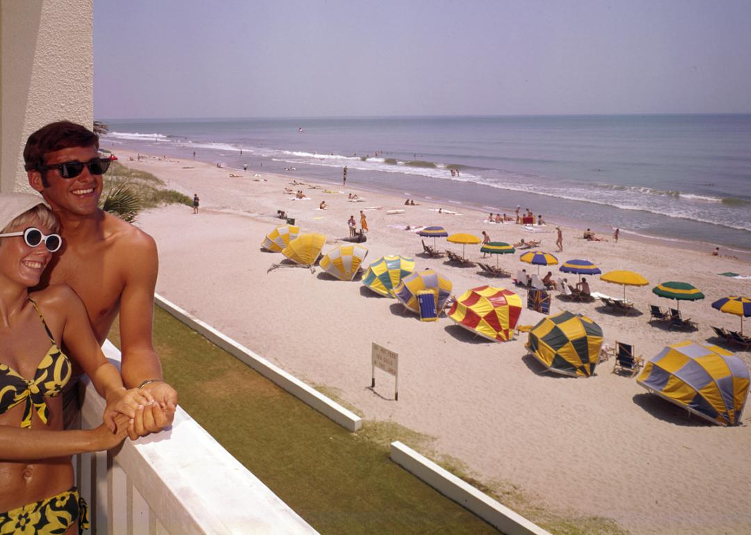 A smiling couple lean on a balcony at Thunderbird Motel, which overlooks a section of beach shaded by various colored umbrellas, Myrtle Beach, South Carolina, 1960s.