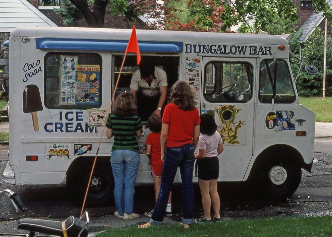 View of a small group of children and teenagers at a Bungalow Bar-brand ice cream truck on Fitchett Street (near 64th Road), in the Rego Park neighborhood, Queens, New York, New York, July 1981.