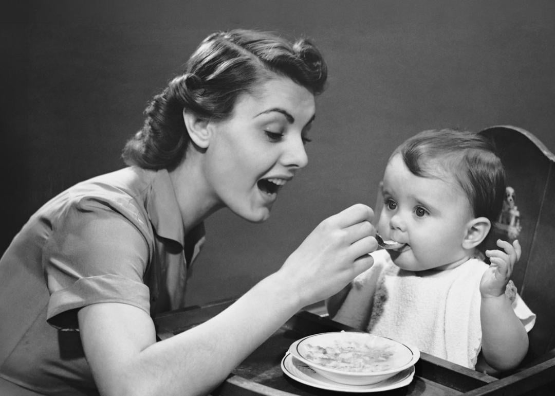 Woman feeding baby in a high chair in the 1950s.