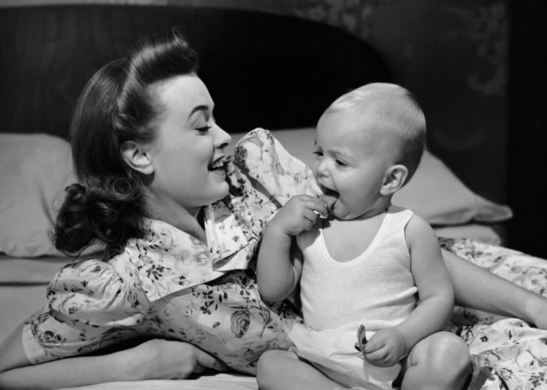 Mother watching her baby eat a cookie, circa 1950s.