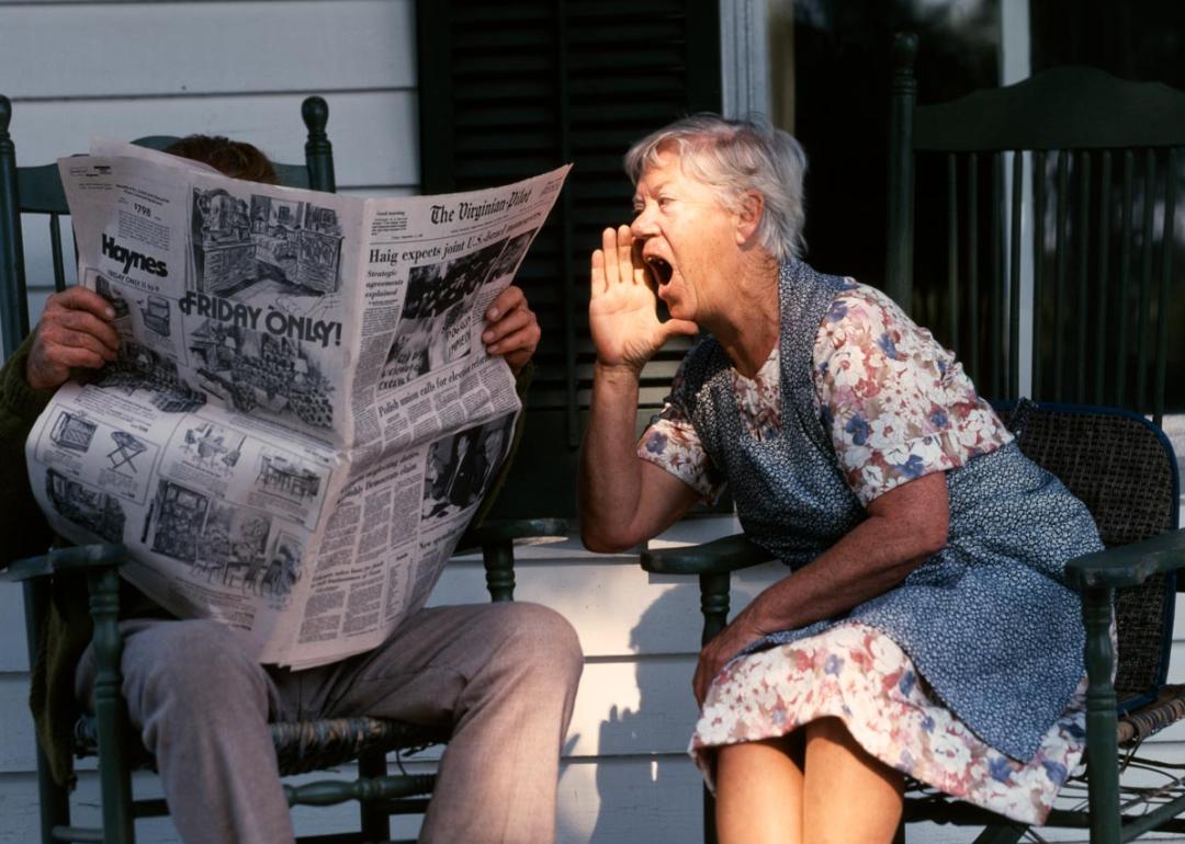 Senior couple sitting on a porch in rocking chairs. Husband is reading the paper and the wife is shouting at him. 