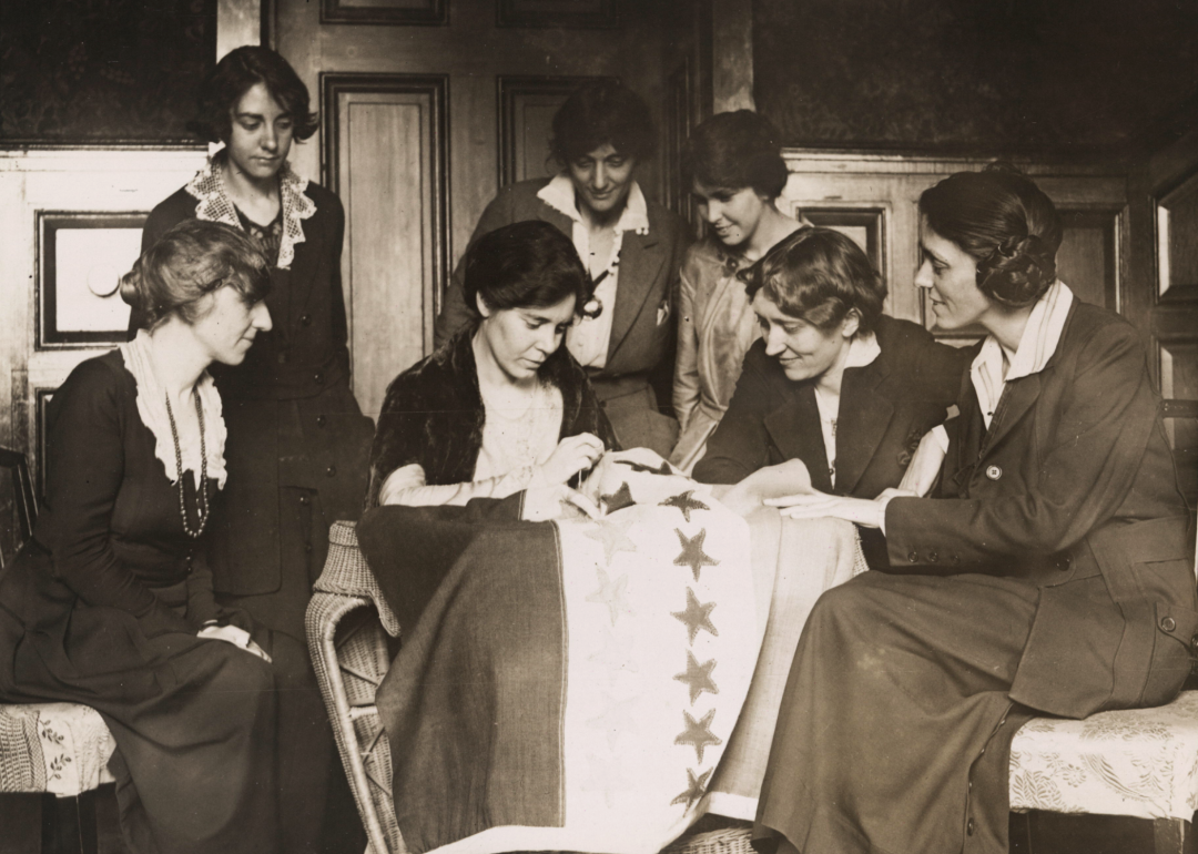 Suffragists watch as Alice Paul sews stars onto banner.