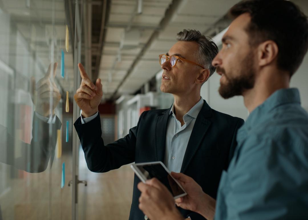 An older man in a suit and glasses standing and pointing to the glass in front of him while the other man is holding a digital screen looking up to the glass.