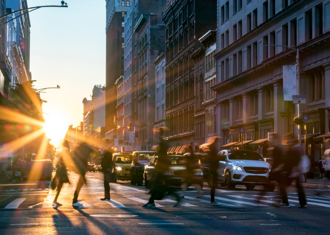 Pedestrians cross a city street at sunset.