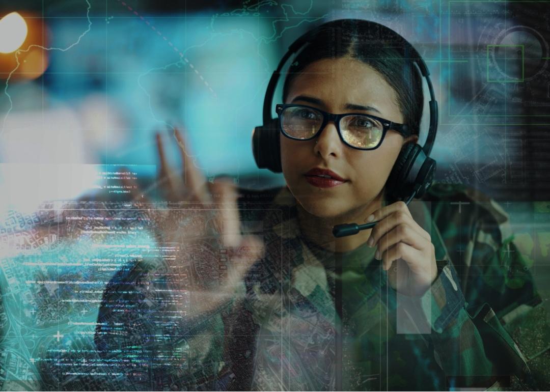 A woman wearing military camouflage and glasses, along with a headset. She is looking on the digital display in front of her.