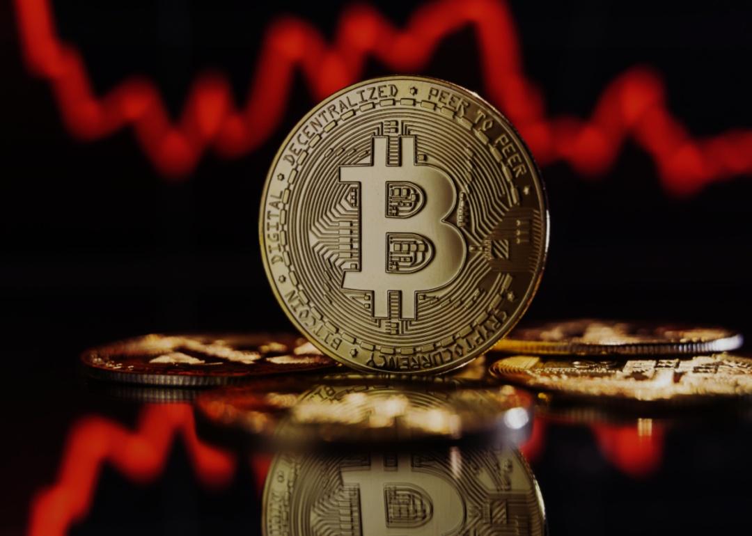 A physical representation of a bitcoin, with the B symbol prominently displayed in the center of the coin, in front of a background of a red, downward-trending line graph.
