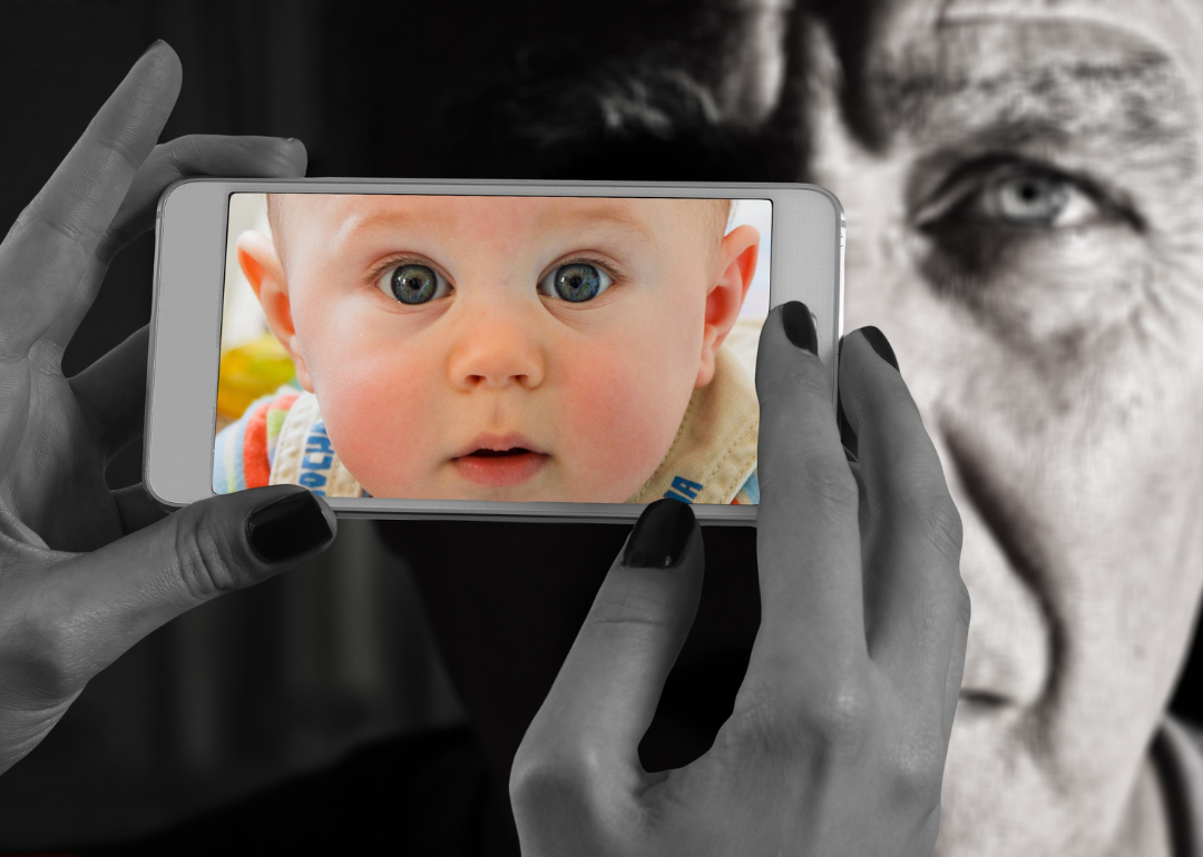 A close up of hands holding a cell phone with an image of a baby on the screen with an older man image in the background. 