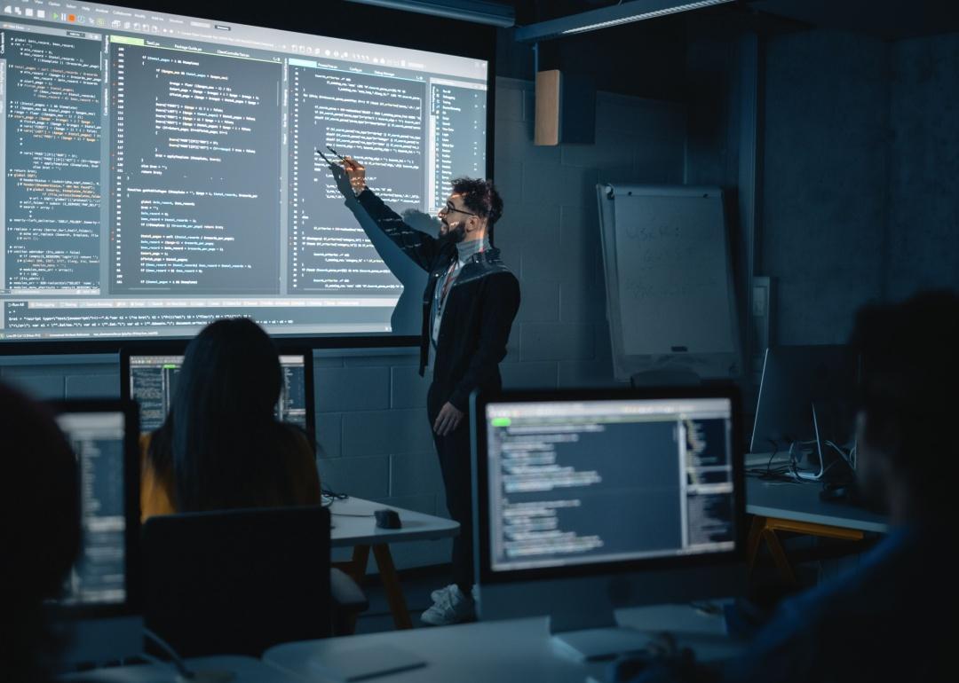 Male teacher pointing at the screen, standing in front of students in a dimly lit classroom.