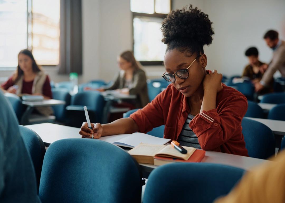 A young woman is seated at a desk. She wears glasses and a casual outfit, with a notebook, pen, and an open book on her desk. In the background, other students are seated in rows.