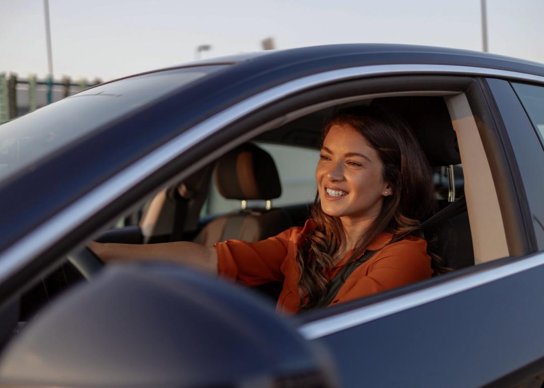 A close up of a young woman sitting in the driver’s seat of a car, smiling warmly.