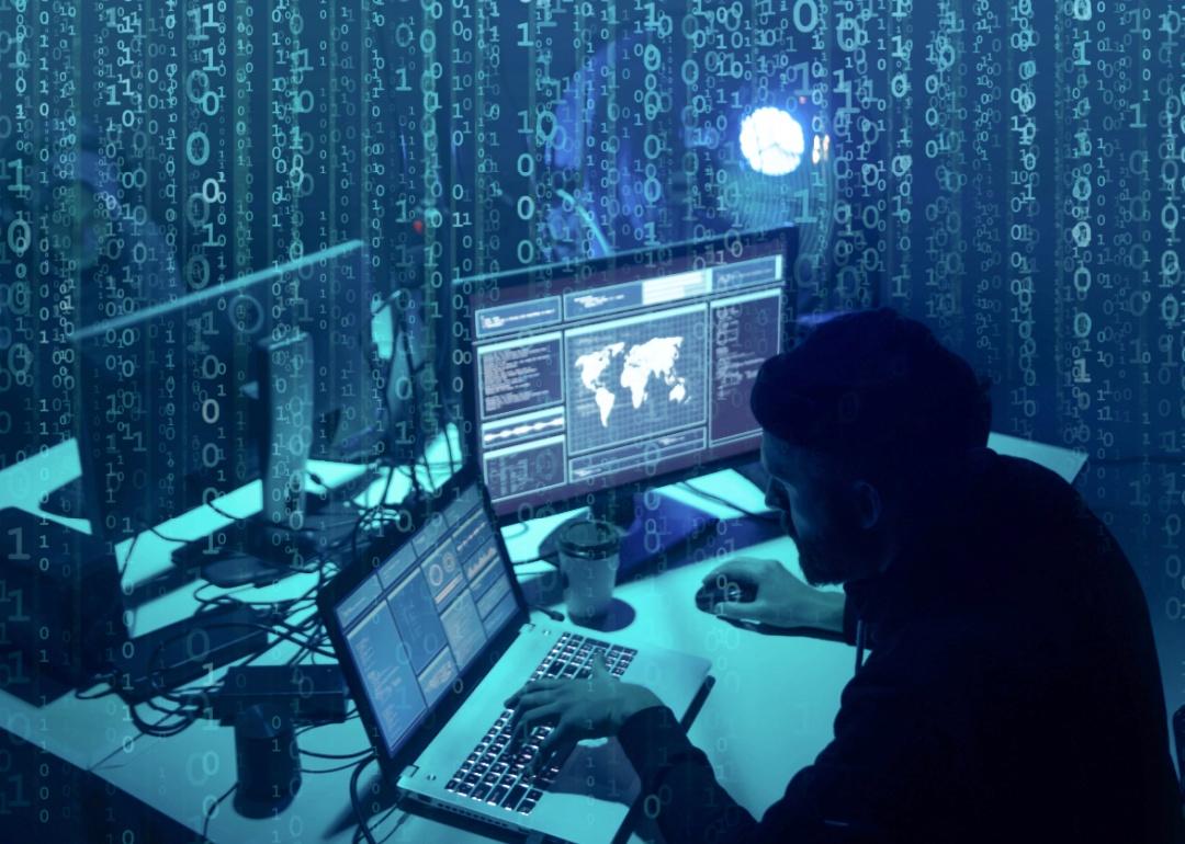 A man's silhouette in a dark room sitting in front of a laptop and another computer screen.