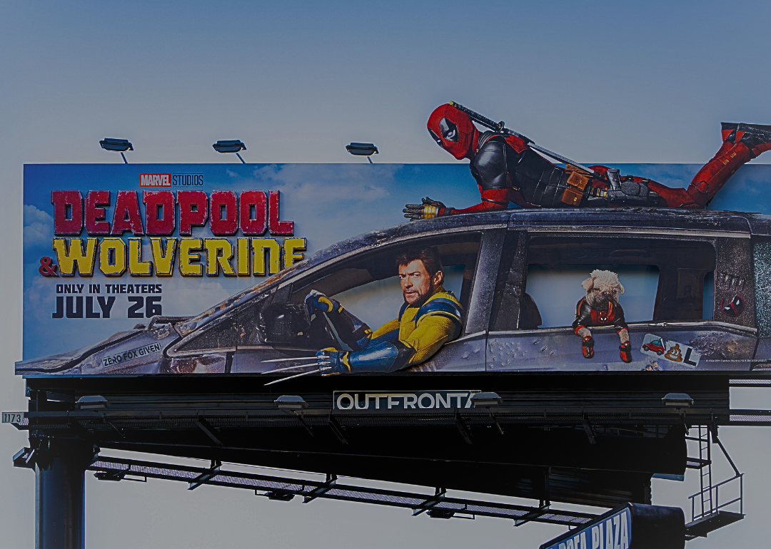 A billboard advertisement promoting the upcoming movie Deadpool & Wolverine by Marvel Studios, set to release on July 26. There is a man dressed in black and red bodysuit lying on top of a damaged car, while a man is in the driver's seat looking focused and intense.