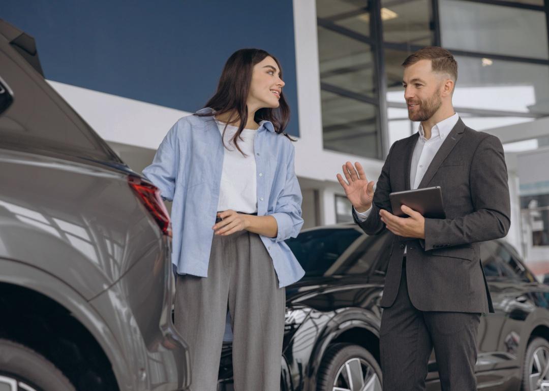 A man dressed in a dark suit holding a tablet is looking towards a young woman in front of him. The woman is casually dressed in a light blue button-down shirt over a white top and gray trousers. She is smiling and appears to be listening to the man next to her. Both individuals are standing next to a car. 