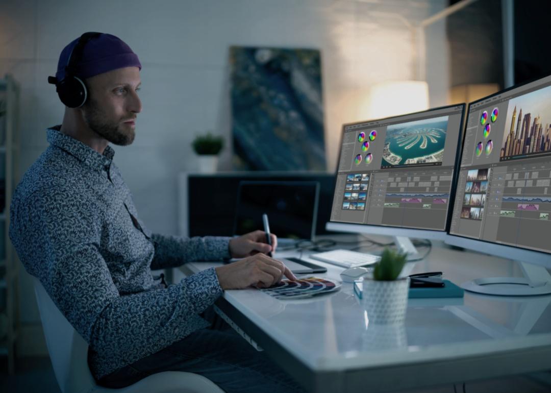 A man wearing a hat and headphones sitting in front of two large computer screens while holding a pen in one hand and pointing to the a color palette with his right index finger.