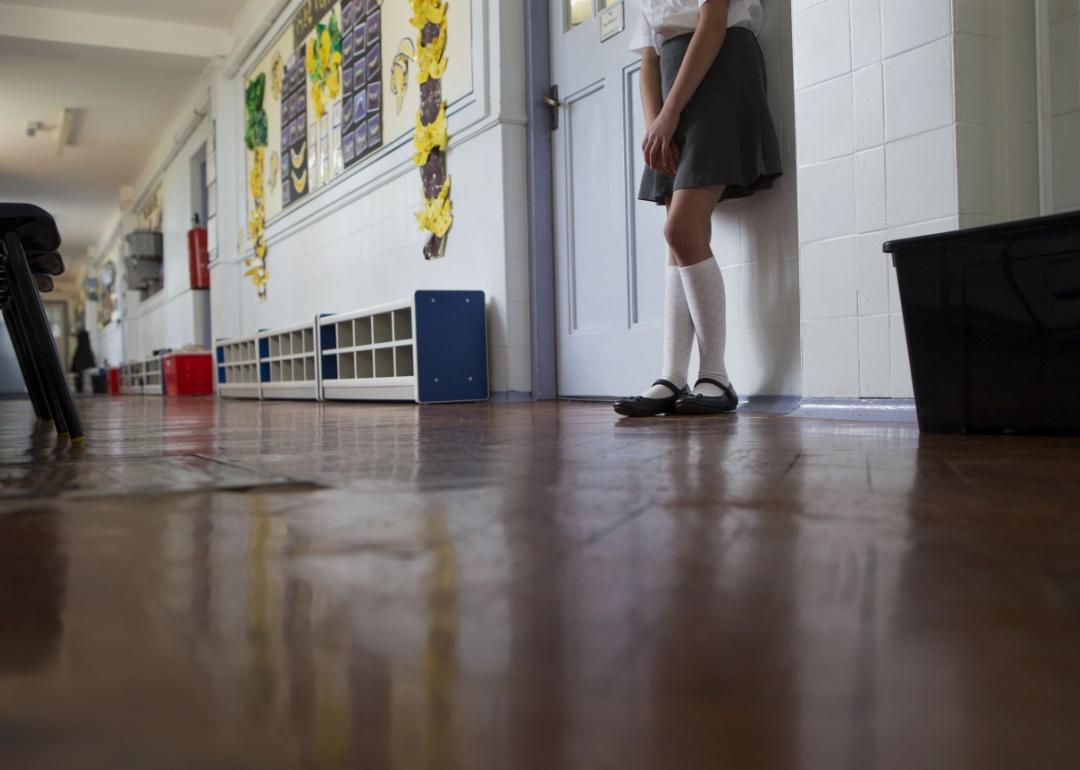 A close-up shot of a young female student's lower torso and legs. She is wearing a grey skirt, long white socks as she leans against a classroom door, standing in a school corridor.