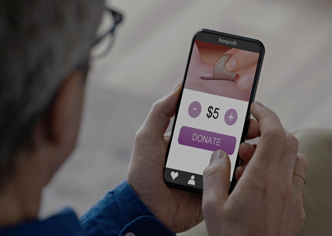 A person holding a smartphone, with a donation application on the screen displaying a coin being dropped into a slot, a selectable donation amount set to "$5" with "+" and "-" buttons and "DONATE" button.