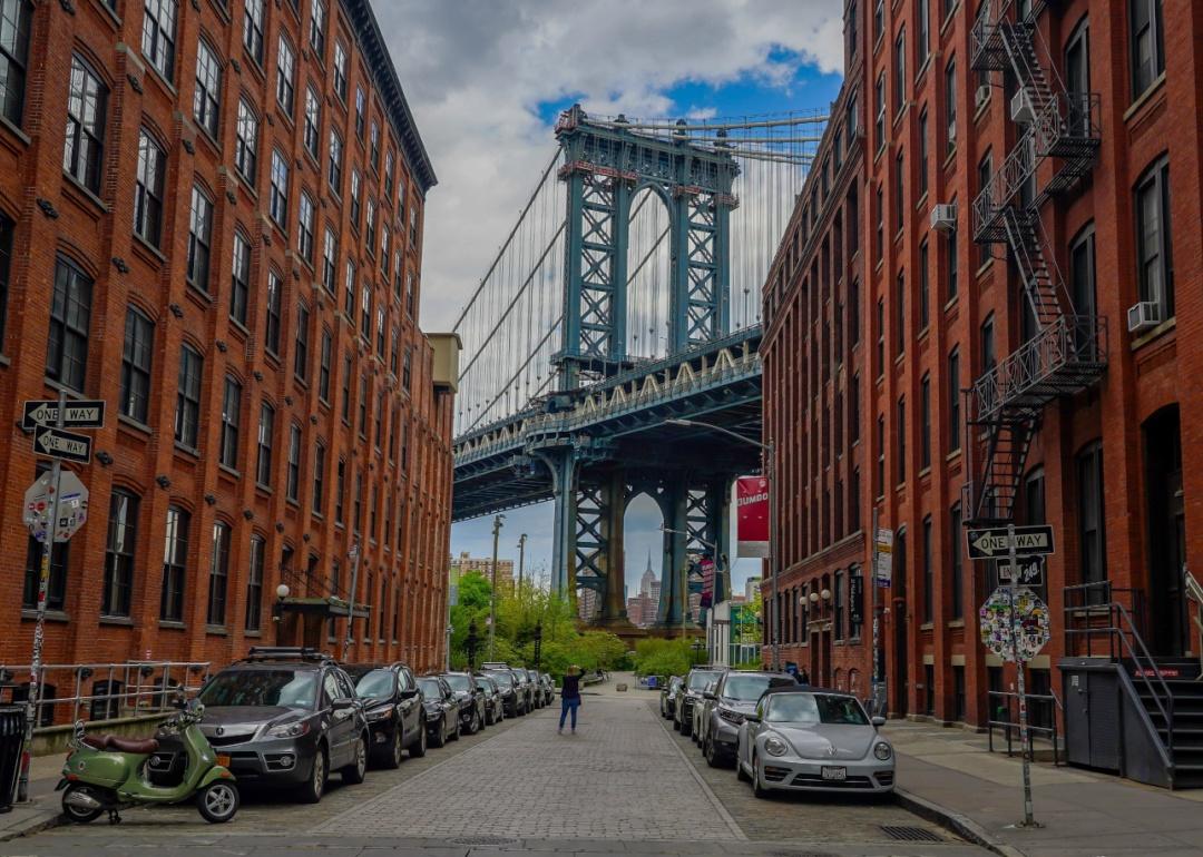 Two rows of red brick industrial buildings with large windows. The cobblestone street is lined with parked cars. An iron bridge is visible in the background.