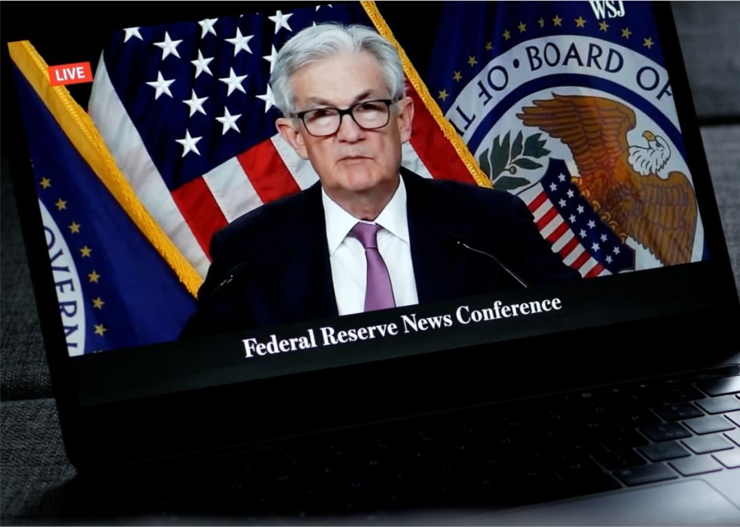 A close-up of a video or broadcast of a "Federal Reserve" news conference on a laptop screen. A man with gray hair is wearing glasses dressed in a suit with a white shirt and purple tie.