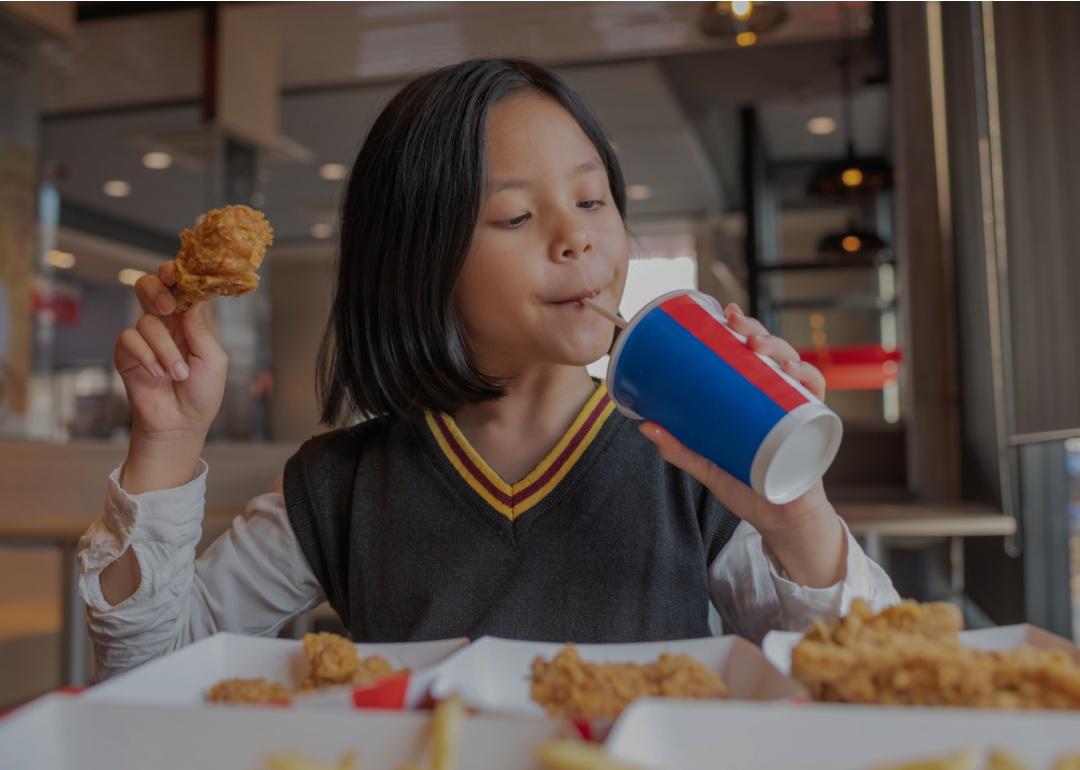A girl sipping a drink and holding a piece of fried chicken sitting in a restaurant. 