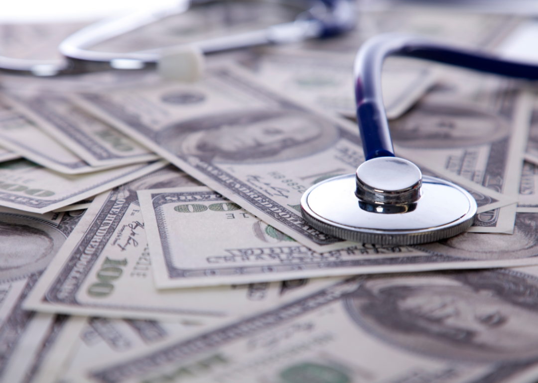 A stethoscope rests on a pile of one hundred dollar bills.