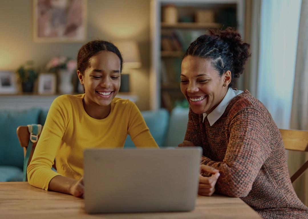 Two people sitting together at a table, both smiling warmly as they look at a laptop screen. The person on the left is a younger African American girl wearing a yellow long-sleeved shirt, looking at the laptop with a bright, happy expression. The person on the right, an African American woman, has her hair styled in a natural updo and is wearing a cozy-looking, textured sweater. 