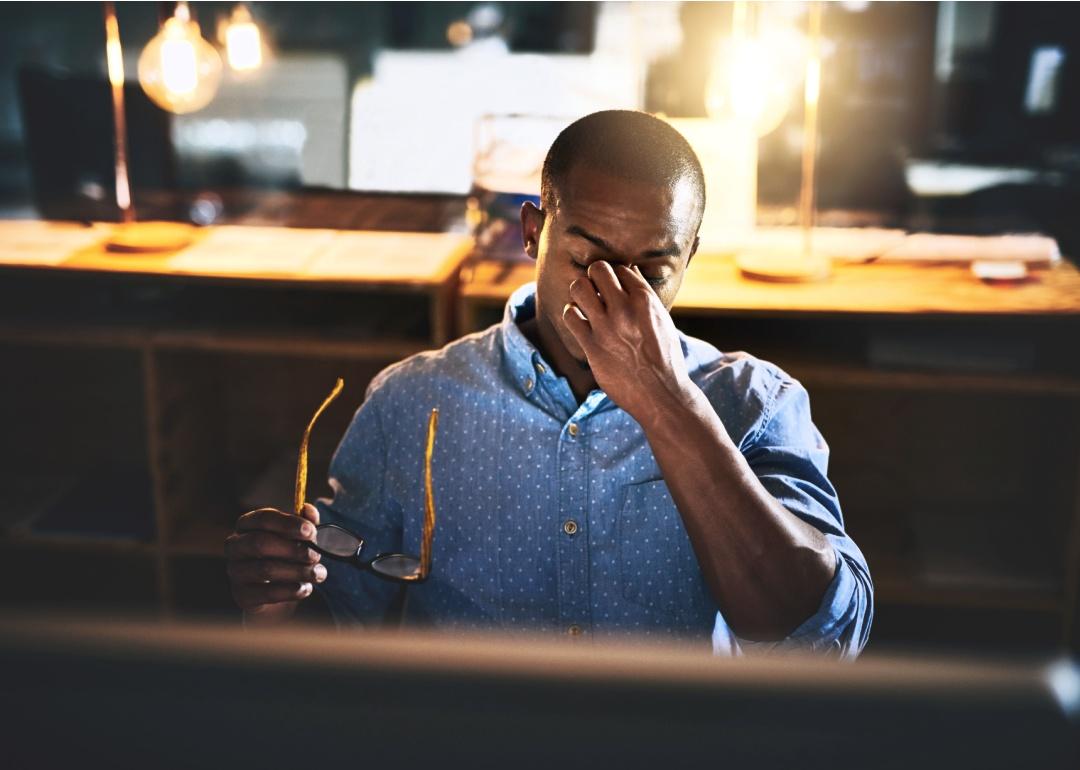 A tired man in a dimly lit workspace sitting at a desk.