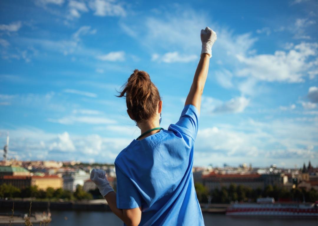 A woman in scrubs holding her hand in the air on a patio facing a city.