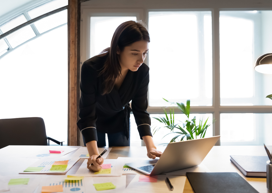 A business person stands at her desk and takes notes while reading her laptop.