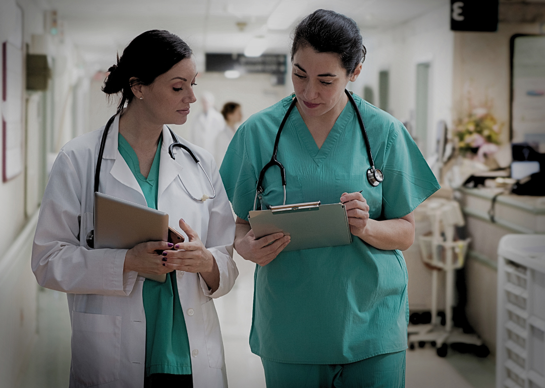 Two women in scrubs walking in a hospital hall looking at the clipboard.  