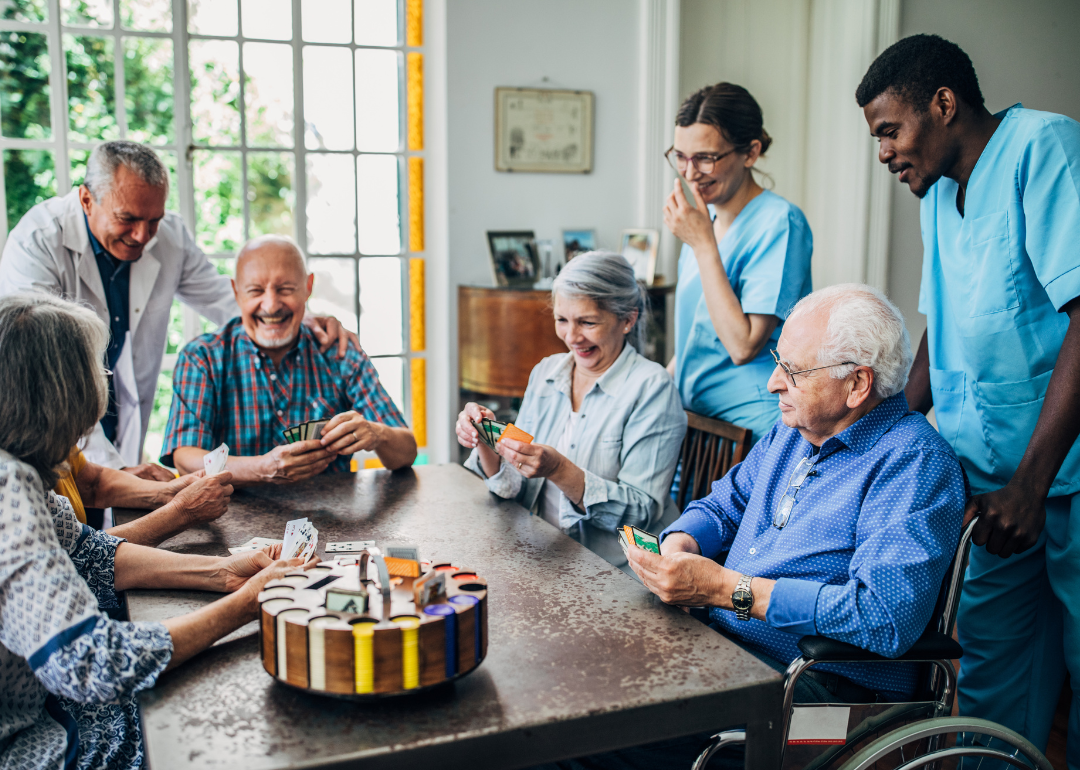 A group of older adults play games at a table with medical staff smiling behind them.