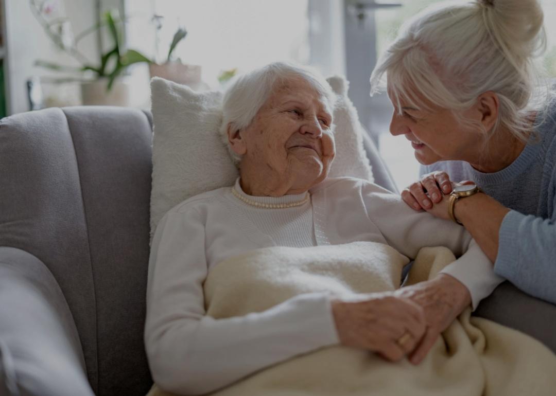 An elderly woman with white hair reclining in a chair, covered with a soft beige blanket. She is wearing a white sweater and a pearl necklace. Beside her, a middle-aged woman leans in holding the elderly woman's hand and smiling.