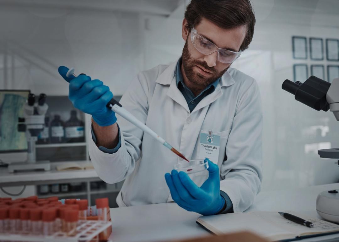 A man in white coat and protective goggles holding a petri dish and a pipette in a lab. 