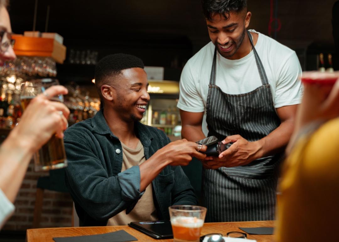 African American man is holdiong his phone close to a credit card reader held by a waiter at a table.