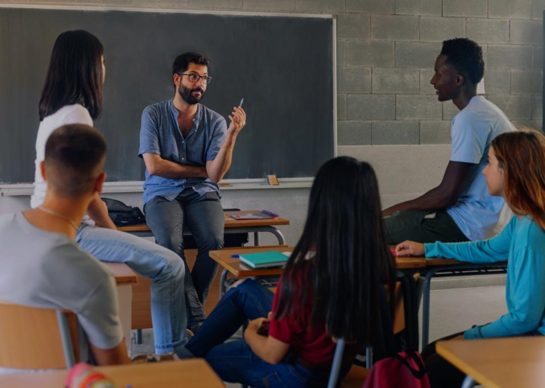 A group of students seated around a teacher in a classroom setting. The teacher casually dressed, is sitting on a desk and appears to be engaged in a discussion with the students. 