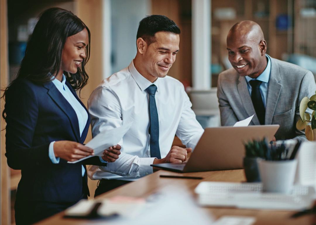 Three smiling businesspeople looking at the laptop on a table in an office.