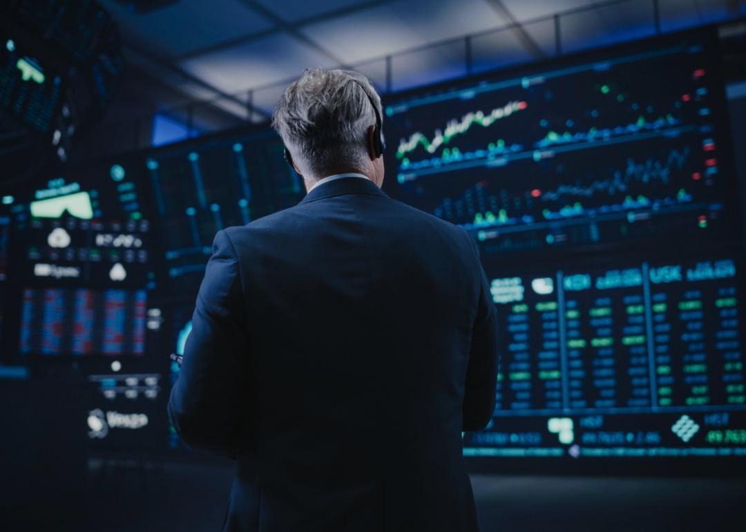 A man standing in front of a large screen displaying multiple charts, graphs, and financial data. The individual is wearing a suit and has a headset on.