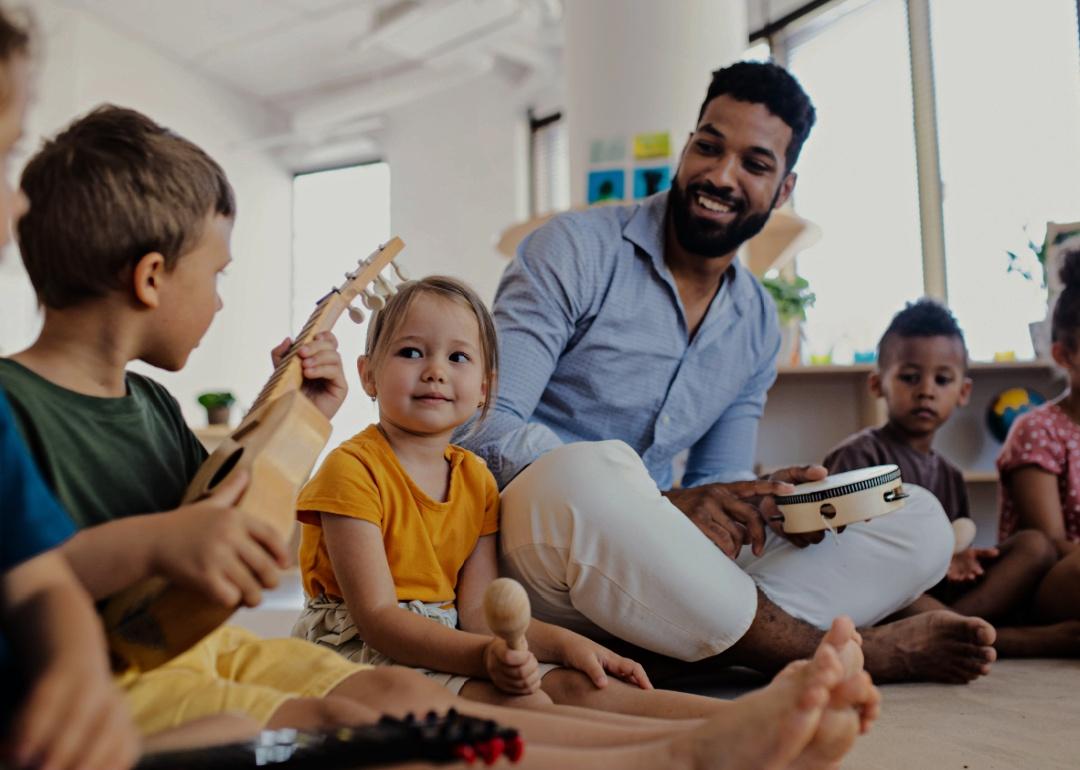 A young male teacher sitting on the floor with a tambourine in his hand surrounded by young kids.