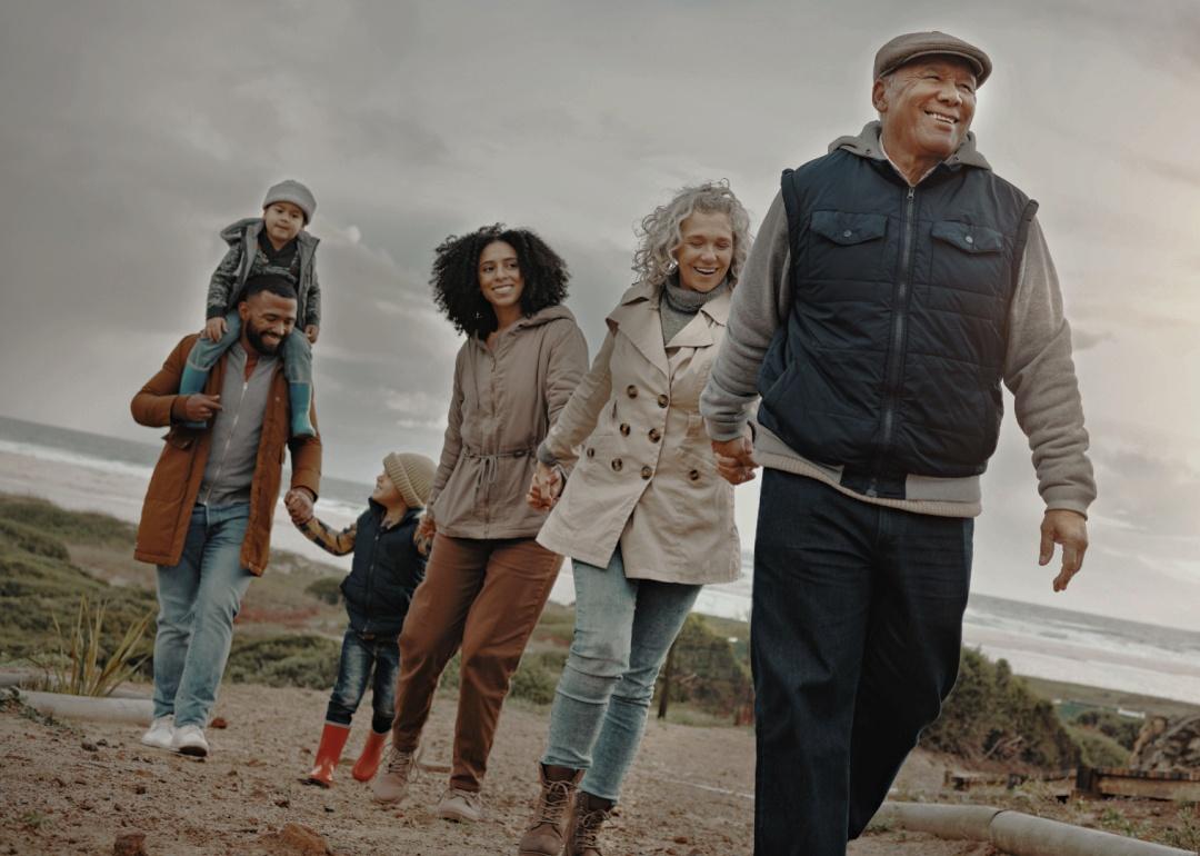 A multigenerational family taking a walk together on a beach. An older man in the front is smiling while holding hands with an older woman and behind them, a younger couple and their children. 