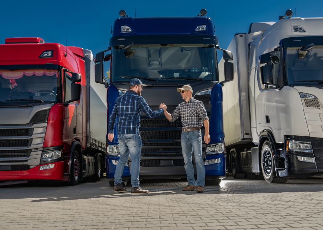 Two men in checked shirts, jeans, and baseball hats shaking hands. There are three truck cabins in the background behind them. 