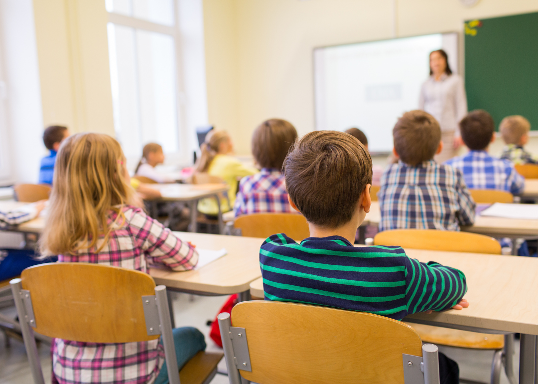 A rear view of young students sitting and listening to teacher in a classroom