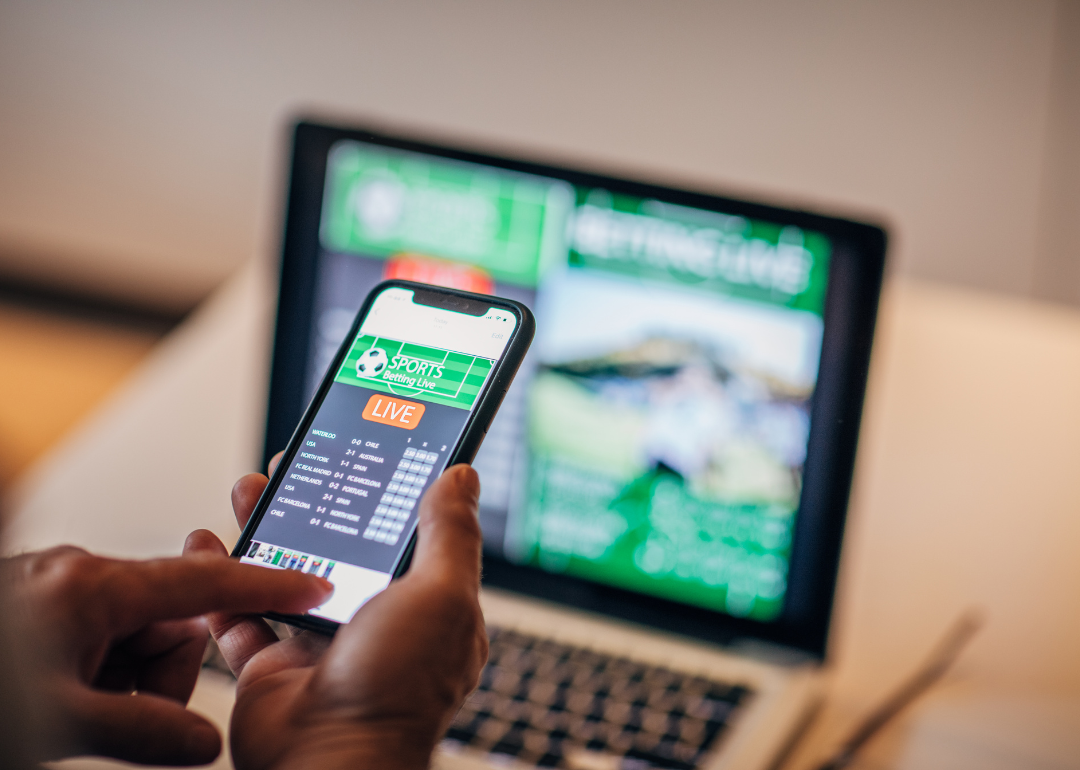 A mobile sports gambling app as seen on a mobile phone in a person's hand and a laptop on a table.