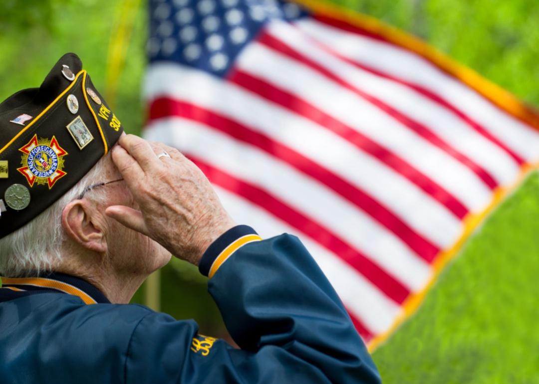 An older veteran saluting with an American flag waving in the background.