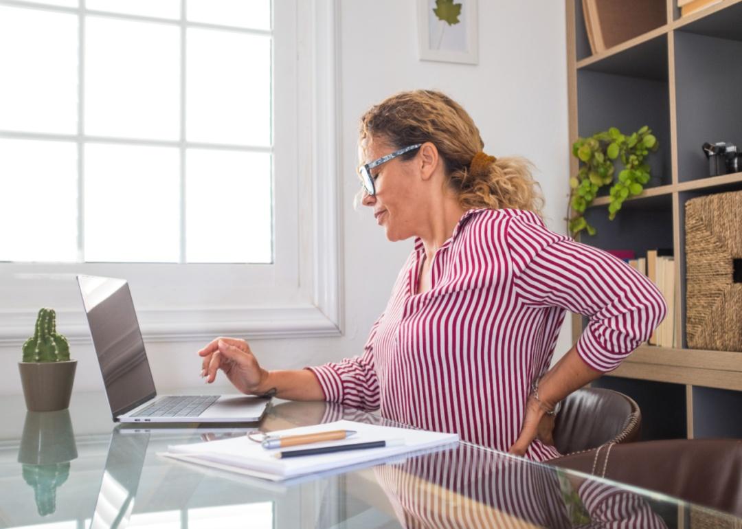 A woman sitting at a desk, grimacing and holding her hand on her back.