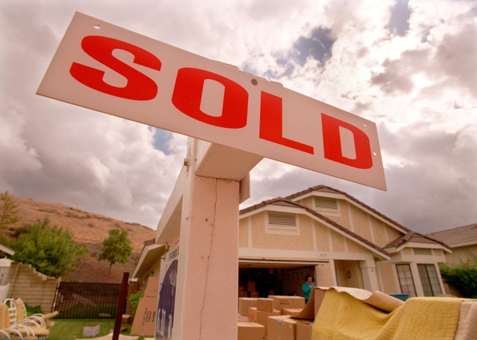 Close-up of a sign reading "Sold" in bright orange in front of a house and lawn full of boxes.